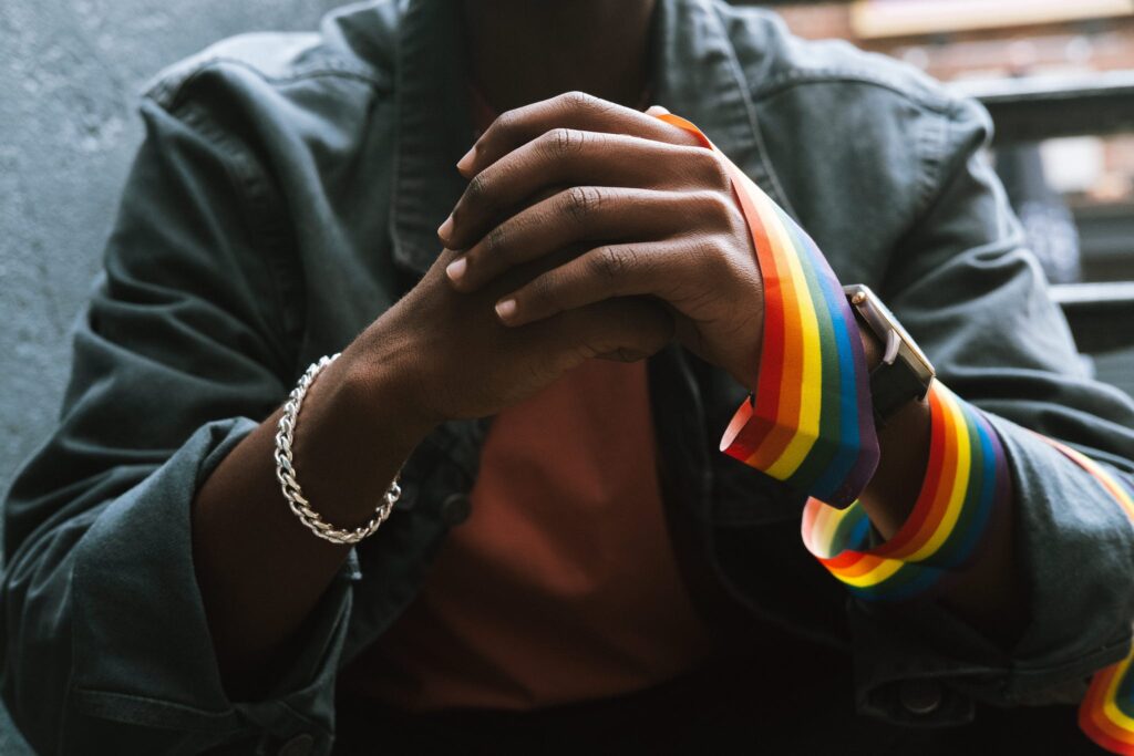 Crop anonymous African American homosexual male in casual clothes wearing LGBT flag on arm while sitting on street and clasping hands